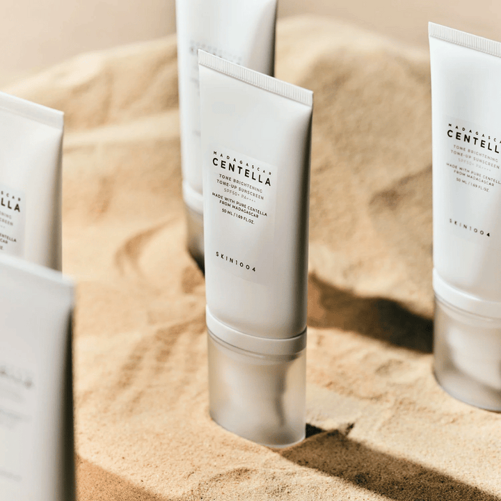 Viaceré tuby s Madagascar Centella Tone Brightening Tone-Up Sunscreen stoja v piesku. Tuba je biela s čiernym textom a obsahuje 50 ml. Produkt má SPF 50+ PA++++ a je vyrobený s čistou Centellou z Madagaskaru.