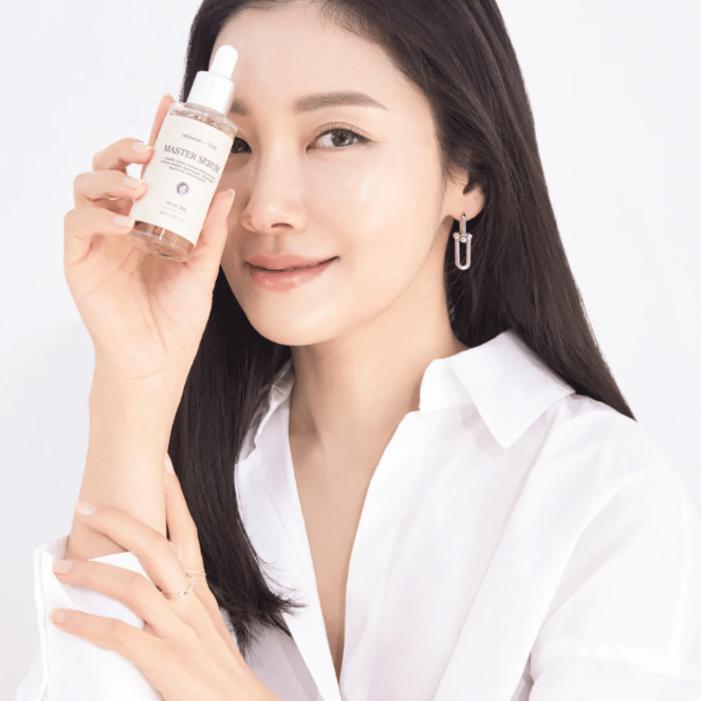 Usmievavá žena s dlhými, tmavými vlasmi a bielou košeľou drží Mixsoon Master Serum vedľa svojej tváre. Jej pleť vyzerá žiarivo a hydratovane.