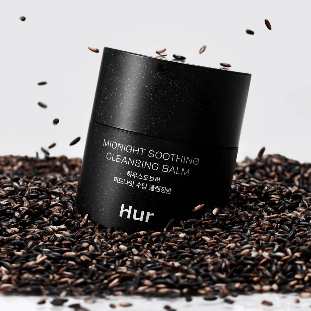 Ako Midnight Soothing Cleansing Balm v čiernej nádobe umiestnenej na čiernych semenách. Upokojujúci čistiaci balzam, ktorý rozpúšťa make-up a nečistoty, zatiaľ čo ošetruje pokožku.