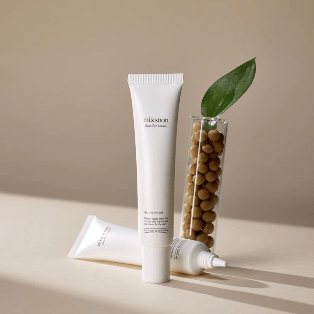 Dve tuby Mixsoon Bean Eye Cream umiestnené vedľa skúmavky naplnenej sójovými bôbmi a zeleným listom rastliny, na béžovom povrchu s hrou tieňov.