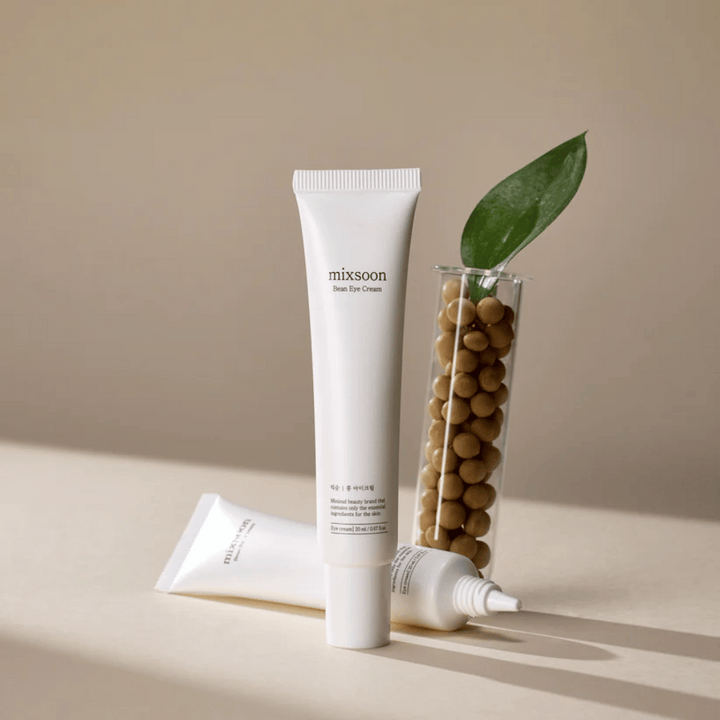Dve tuby Mixsoon Bean Eye Cream umiestnené vedľa skúmavky naplnenej sójovými bôbmi a zeleným listom rastliny, na béžovom povrchu s hrou tieňov.