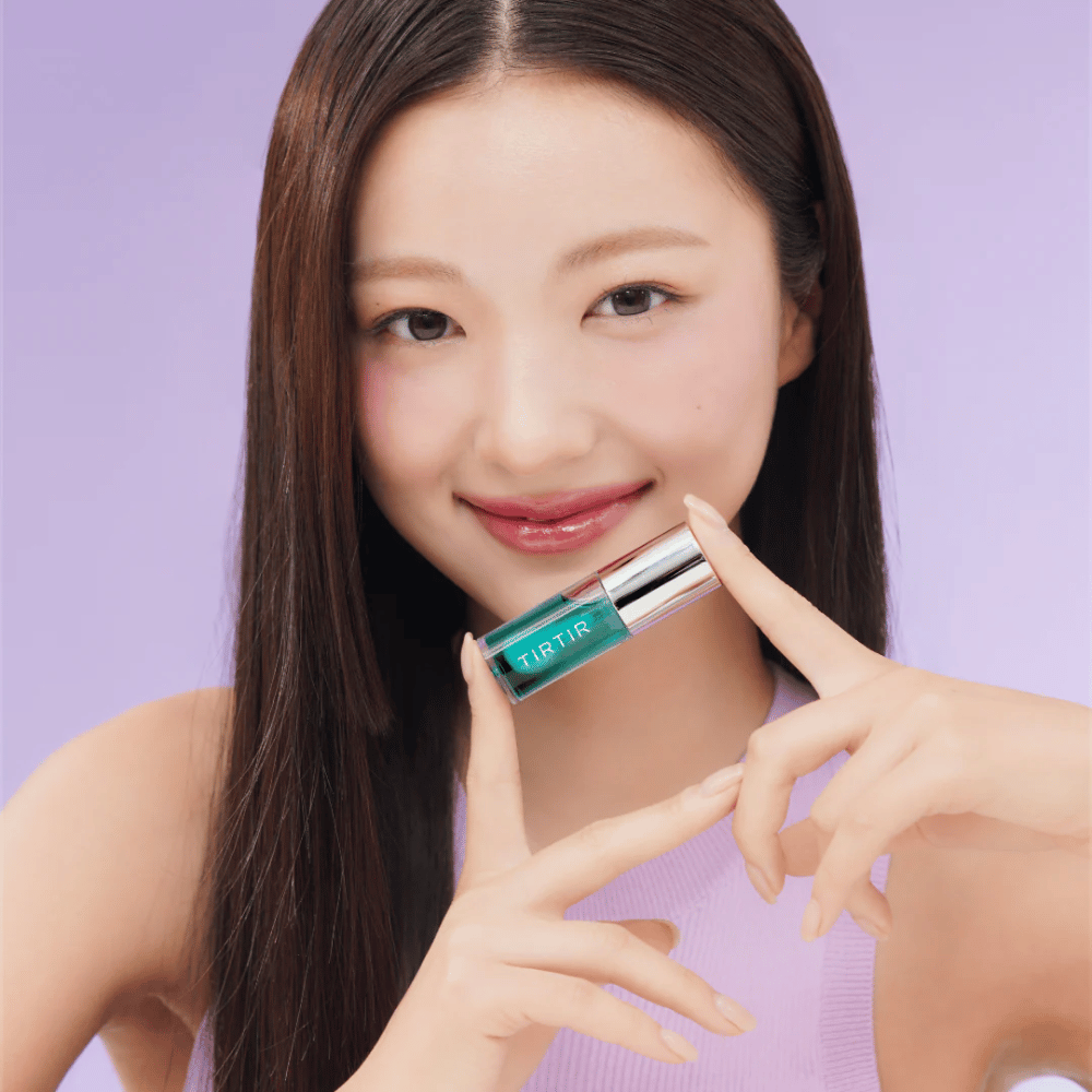 Žena s dlhými, rovným vlasmi drží TIRTIR My Glow Lip Oil a ukazuje svoje lesklé, ružové pery na fialovom pozadí.