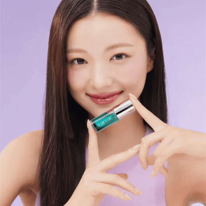 Žena s dlhými, rovným vlasmi drží TIRTIR My Glow Lip Oil a ukazuje svoje lesklé, ružové pery na fialovom pozadí.