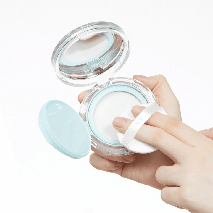 Ruka drží otvorenú nádobu s VT PDRN Essence Glow Sun Pact. Puff sa chystá aplikovať púder, ktorý má biely povrch a modrý okraj okolo nádoby.