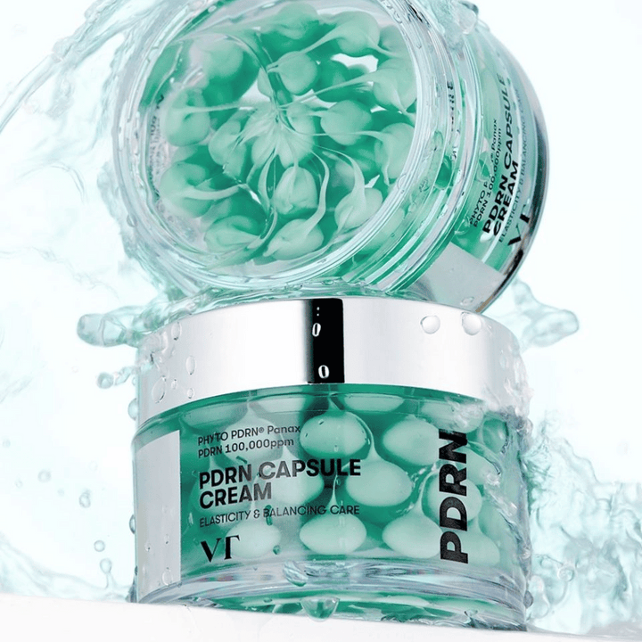 Dve nádoby VT PDRN Capsule Cream naskladané s rozstrekmi vody okolo. Priehľadné nádoby ukazujú zelené kapsuly vo vnútri, s textom "Elasticity & Balancing Care" viditeľným na etikete.