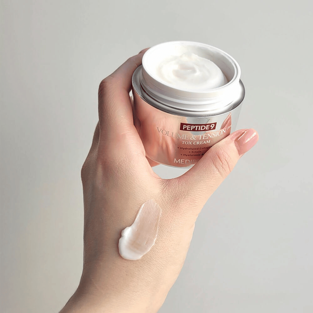 Ruka drží MEDI-PEEL Peptide 9 Volume & Tension Tox Cream s kvapkou krému na chrbte ruky. Hodvábna formula hydratuje a dodáva pokožke pevnosť a pružnosť.
