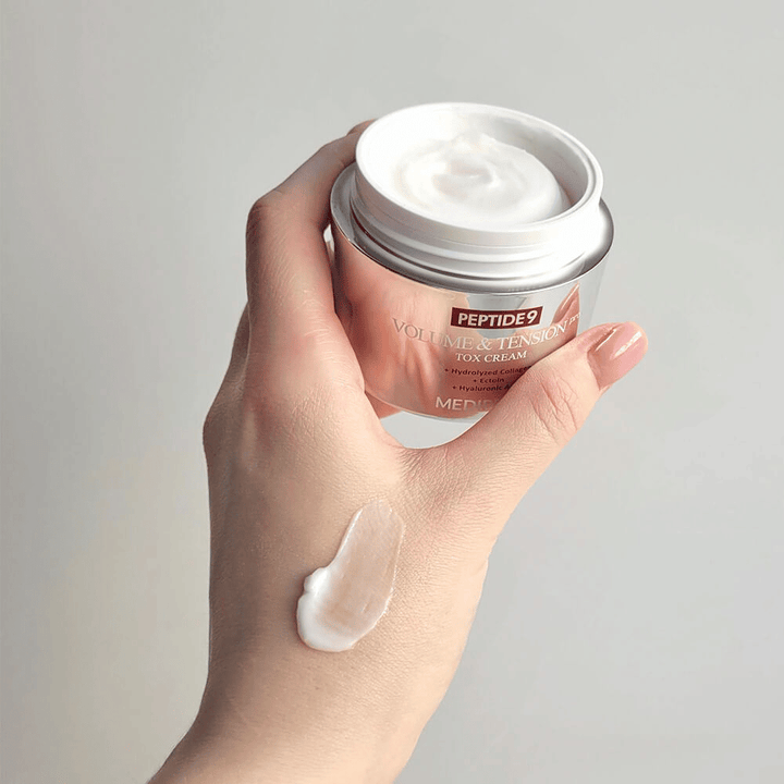 Ruka drží MEDI-PEEL Peptide 9 Volume & Tension Tox Cream s kvapkou krému na chrbte ruky. Hodvábna formula hydratuje a dodáva pokožke pevnosť a pružnosť.