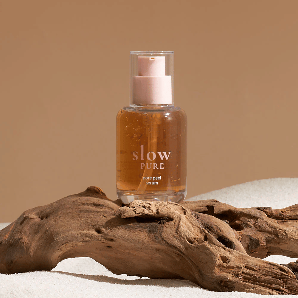 Slow Pure Pore Peel Serum umiestnené na piesku s dreveným detailom v pozadí, symbol pre prírodnú starostlivosť o pleť.