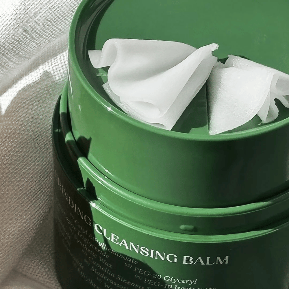 Obrázok ukazuje zelenú nádobu s "Cleansing Balm", ktorá je otvorená, aby odhalila priloženú špachtľu a jemný obsah. Krémová konzistencia odráža svetlo, čo vyvoláva pocit luxusu a efektívnosti pri čistení.