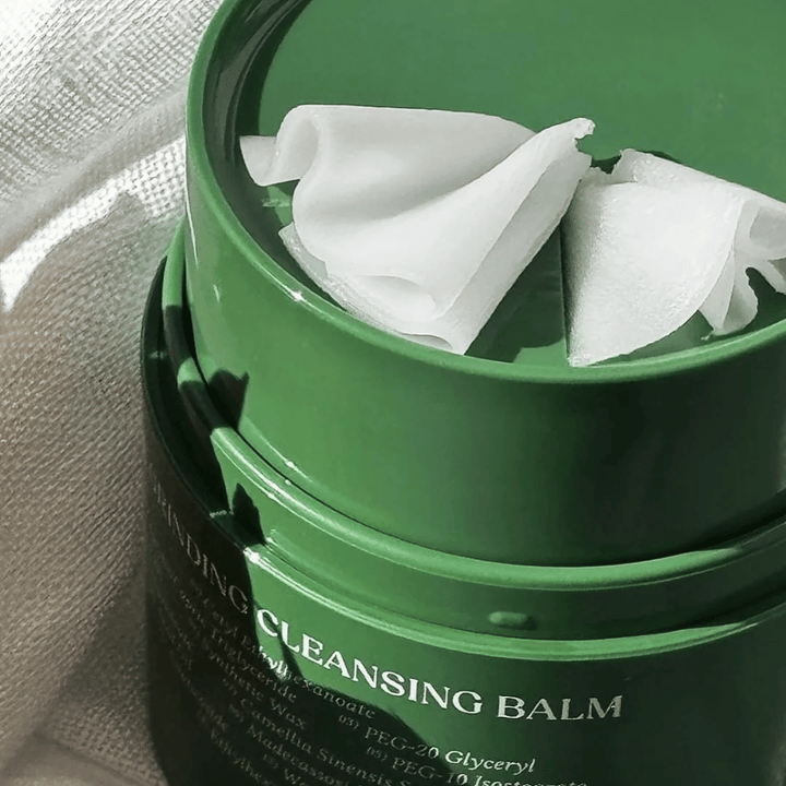 Obrázok ukazuje zelenú nádobu s "Cleansing Balm", ktorá je otvorená, aby odhalila priloženú špachtľu a jemný obsah. Krémová konzistencia odráža svetlo, čo vyvoláva pocit luxusu a efektívnosti pri čistení.