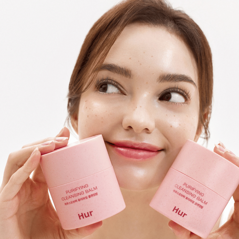 Modelka drží dve nádoby Ako Purifying Cleansing Balm. Jemný čistiaci balzam, ktorý je vhodný pre citlivú pokožku a poskytuje svieži, hydratovaný pocit po čistení.