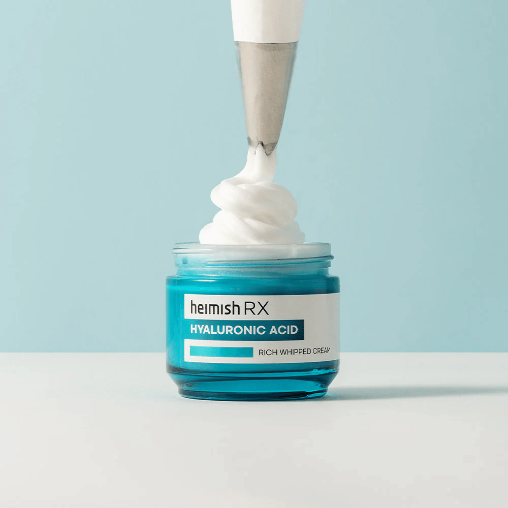 Heimish RX Hyaluronic Acid Rich Whipped Cream v modrej sklenenej nádobe, kde cukrárska taška nanáša nadýchaný krém. Hydratačný pleťový krém s kyselinou hyalurónovou pre intenzívnu hydratáciu a jemnú, plnú pokožku.
