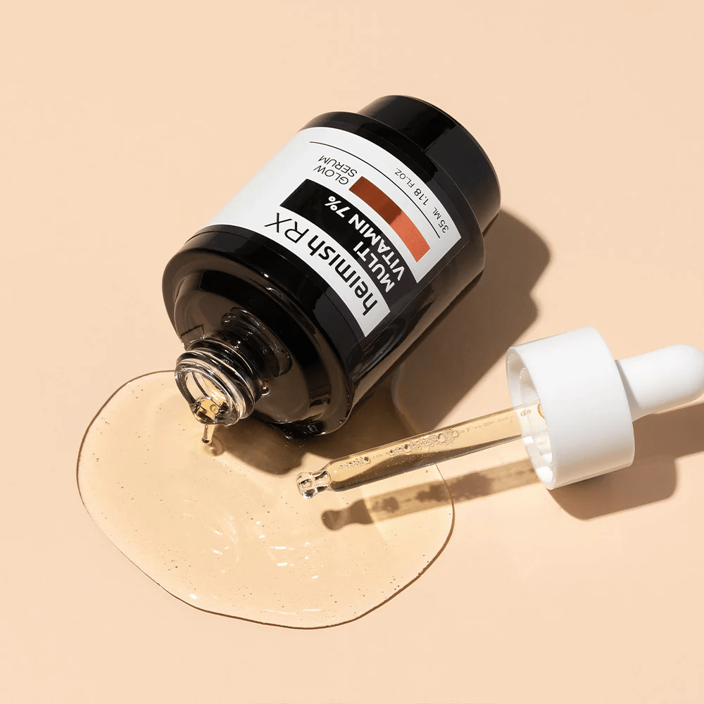 Heimish RX Multi Vitamin 7% Glow Serum ležiace na béžovom pozadí so sérom rozliatym okolo fľaštičky a pipetou naplnenou produktom, čo zdôrazňuje jeho výživnú formulu.