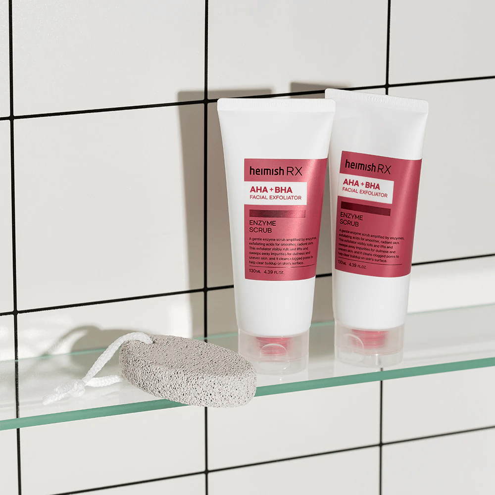 Dve tuby Heimish RX AHA + BHA Facial Exfoliator umiestnené na sklenenej poličke v kúpeľni s bielou kachličkovou stenou. Vedľa produktov leží pemza s bielym šnúrkou. Elegantný a minimalistický obraz starostlivosti o pleť.