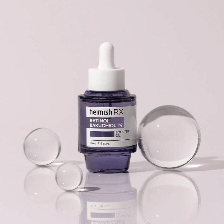 Heimish RX Retinol Bakuchiol Booster Oil 1% v elegantnej fialovej sklenenej fľaštičke s pipetou. Olej je umiestnený na reflexnom povrchu s okolo rozmiestnenými sklenenými guľôčkami, čo vytvára luxusný a moderný pocit.