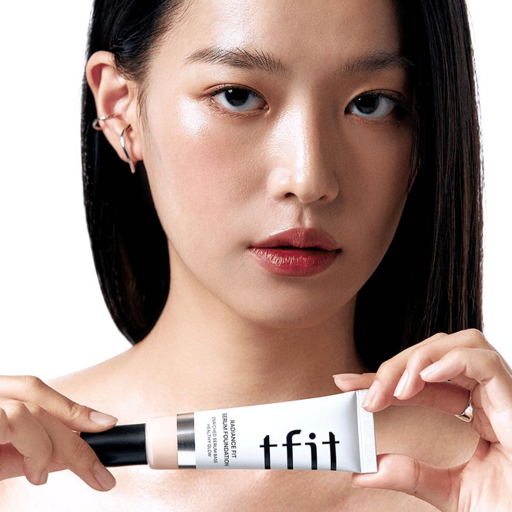 Modelka s lesklou, hladkou pokožkou drží TFIT Radiance Fit Serum Foundation, čo zvýrazňuje prirodzený finiš a lesk produktu.