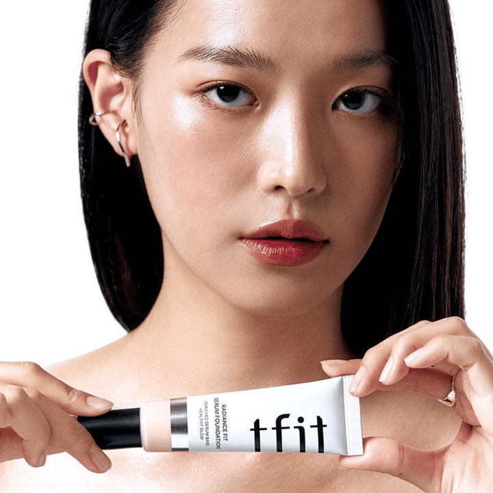 Modelka s lesklou, hladkou pokožkou drží TFIT Radiance Fit Serum Foundation, čo zvýrazňuje prirodzený finiš a lesk produktu.