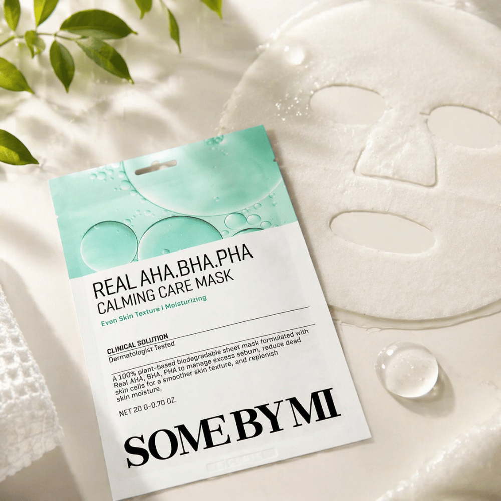 SOME BY MI Real AHA.BHA.PHA Calming Care Mask je zobrazená spolu s rozvinutou plátennou maskou. Okolo produktu sú viditeľné kvapky vlhkosti a zelené listy, ktoré posilňujú pocit hydratácie, upokojujúcej starostlivosti a jemnej exfoliácie. Obrázok zdôrazňuje zameranie masky na rovnomernejšiu textúru pleti a zlepšenú rovnováhu hydratácie.