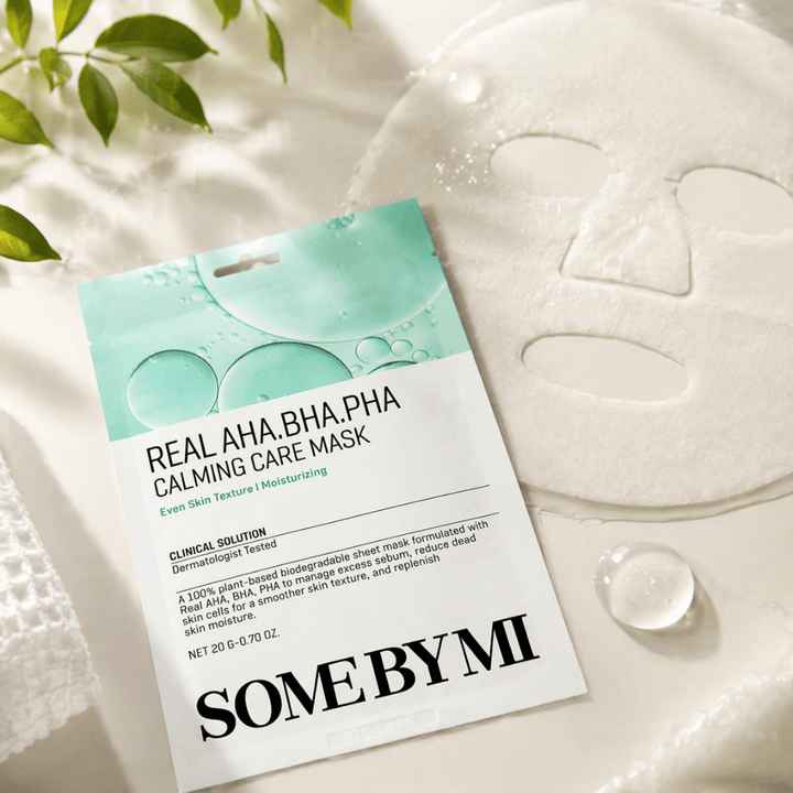 SOME BY MI Real AHA.BHA.PHA Calming Care Mask je zobrazená spolu s rozvinutou plátennou maskou. Okolo produktu sú viditeľné kvapky vlhkosti a zelené listy, ktoré posilňujú pocit hydratácie, upokojujúcej starostlivosti a jemnej exfoliácie. Obrázok zdôrazňuje zameranie masky na rovnomernejšiu textúru pleti a zlepšenú rovnováhu hydratácie.