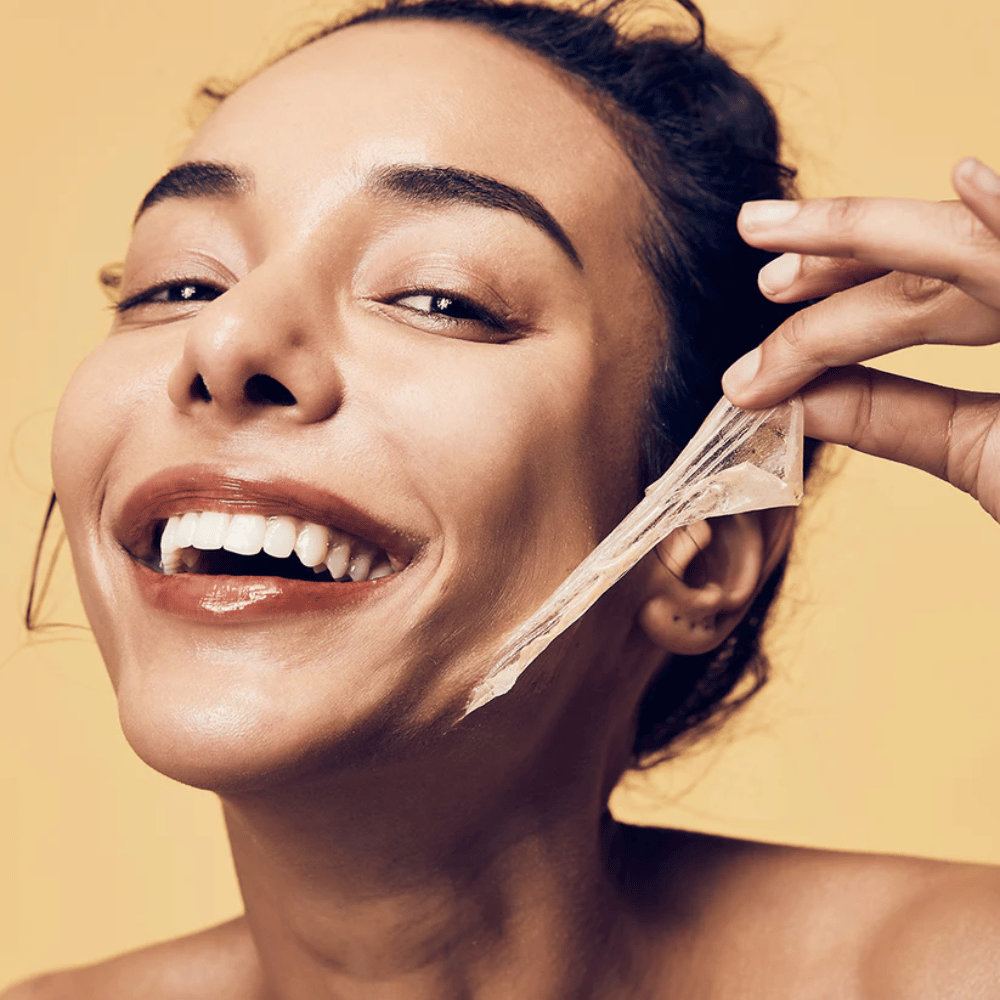 Obrázok ukazuje ženu s veľkým úsmevom, ktorá si práve odstraňuje peel-off masku z tváre. Žena vyzerá šťastne a spokojne, keď používa kozmetický produkt, čo sprostredkúva pocit pohody a starostlivosti o pleť.