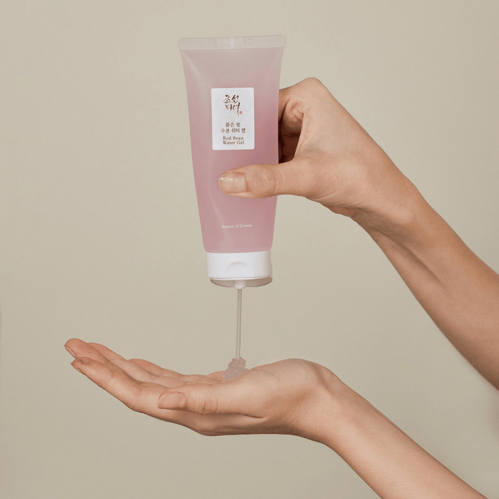 Obrázok zobrazuje osobu, ktorá drží tubu "Red Bean Water Gel" od Beauty of Joseon a vytláča priehľadný gél do svojej ruky. Produkt vyzerá byť určený na starostlivosť o pleť a má atraktívne, jemne ružové balenie.