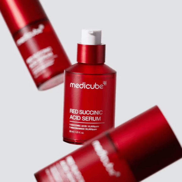 Medicube Red Succinic Acid Serum v červenej pumpovacej fľaši, obklopené viacerými identickými fľašami na svetlom pozadí. Produkt obsahuje kyselinu jantárovú a niacínamid na liečbu akné a pigmentových škvŕn.
