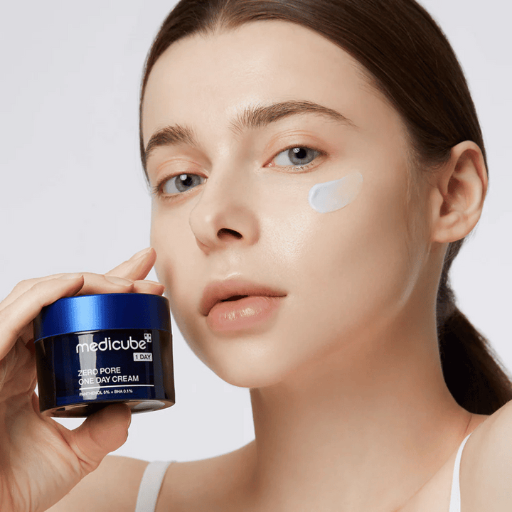 Žena s prirodzeným vzhľadom drží Medicube Zero Pore One Day Cream. Malé množstvo krému je nanesené na jej líci. Produkt je navrhnutý na zlepšenie štruktúry pokožky a minimalizáciu veľkosti pórov, s ľahkou a hydratačnou formulou.