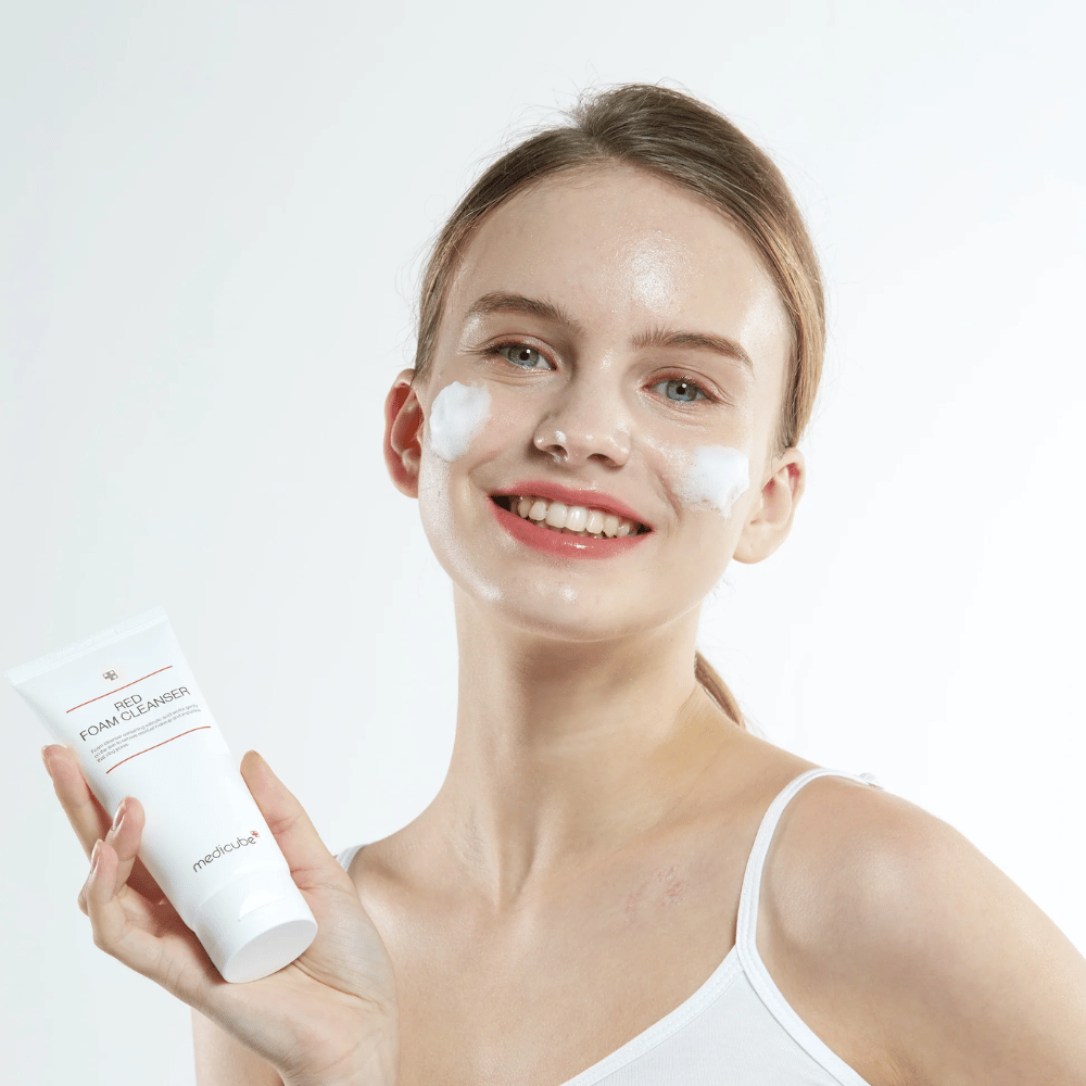 Mladá žena s čistiacou penou na tvári drží tubu Medicube Red Foam Cleanser a usmieva sa do kamery. Má na sebe biele tielko a pozadie je svetlé a jednoduché.