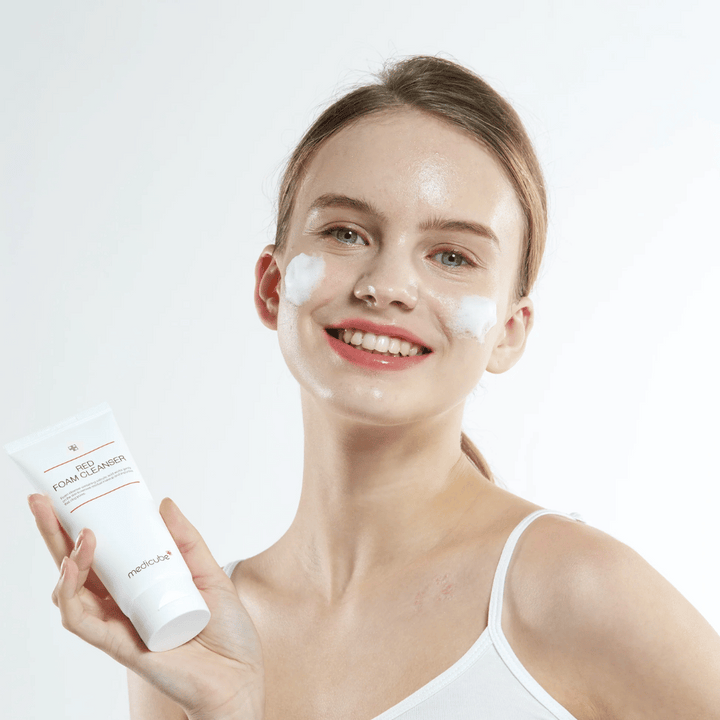 Mladá žena s čistiacou penou na tvári drží tubu Medicube Red Foam Cleanser a usmieva sa do kamery. Má na sebe biele tielko a pozadie je svetlé a jednoduché.
