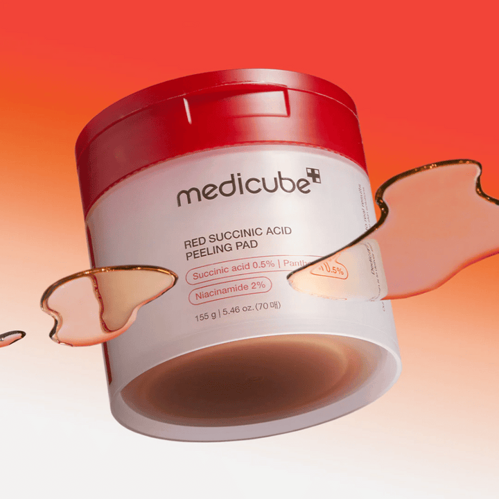 Medicube Red Succinic Acid Peeling Pad v bielej a červenej nádobe, umiestnená na pozadí s prechodom červenej a oranžovej farby. Produkt obsahuje kyselinu jantárovú a niacínamid na exfoliáciu a zlepšenie pokožky. Tekutá textúra je viditeľná okolo nádoby.