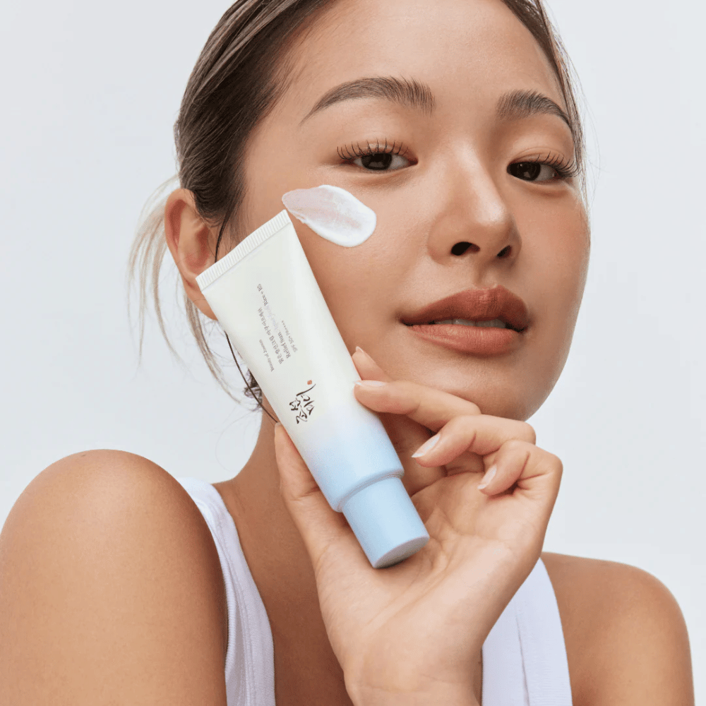 Žena drží Beauty of Joseon Relief Sun: Aqua-Fresh Rice + B5 opaľovací krém a ukazuje jeho krémovú konzistenciu na líci. Ľahký opaľovací produkt, ktorý hydratuje a chráni pokožku s SPF 50+ PA++++.