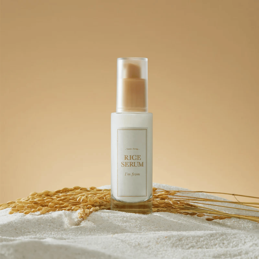 Fľaša s "I'm from" Rice Serum, obklopená ryžovými zrnami a ryžovými klasmi na teplom pozadí.