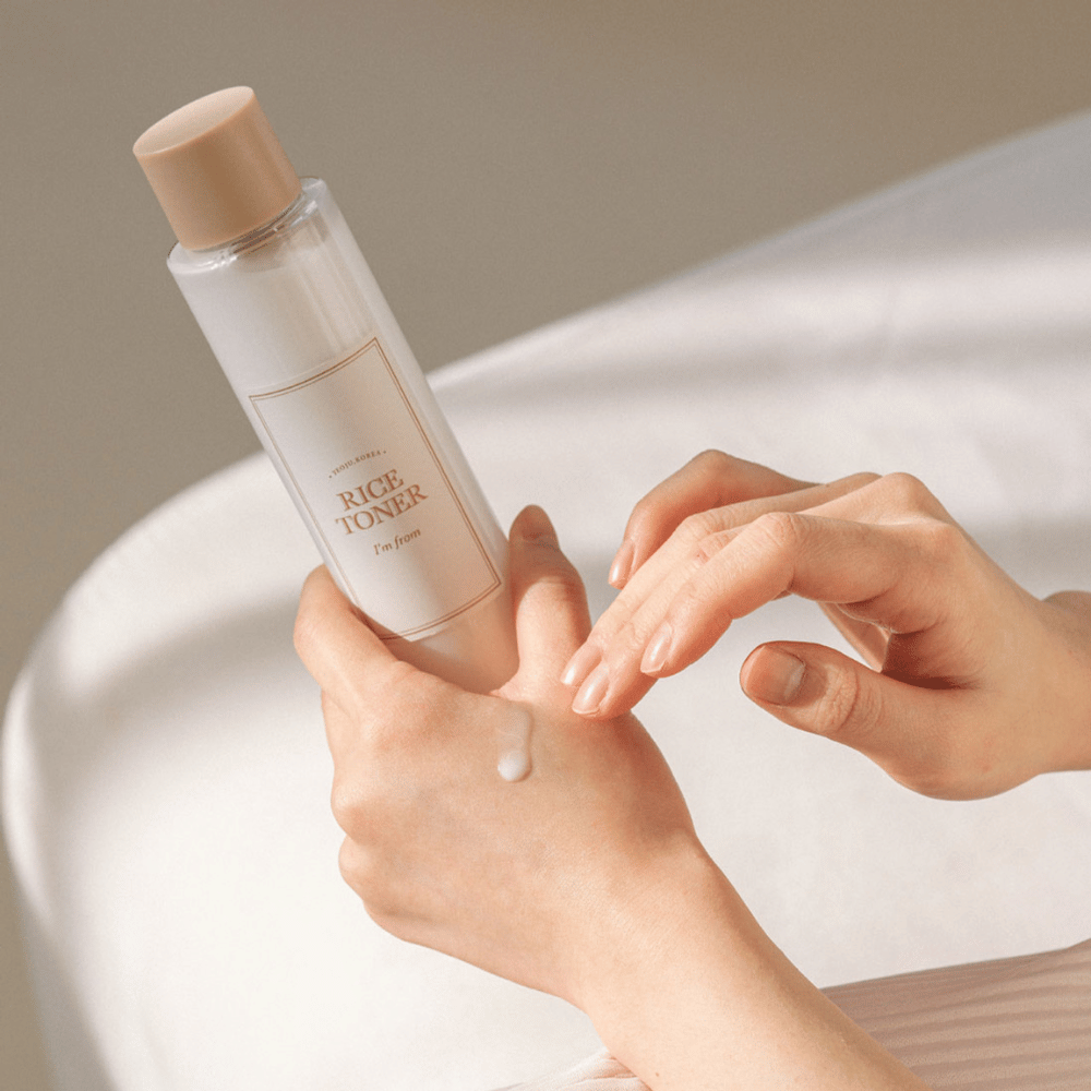 Ruky držia fľašu ryžového tonera označenú "I'm from". Kvapka produktu je aplikovaná na jednu ruku, zatiaľ čo druhá ruka sa pripravuje na jej aplikáciu. Scéna je jemne osvetlená s teplým tónom.