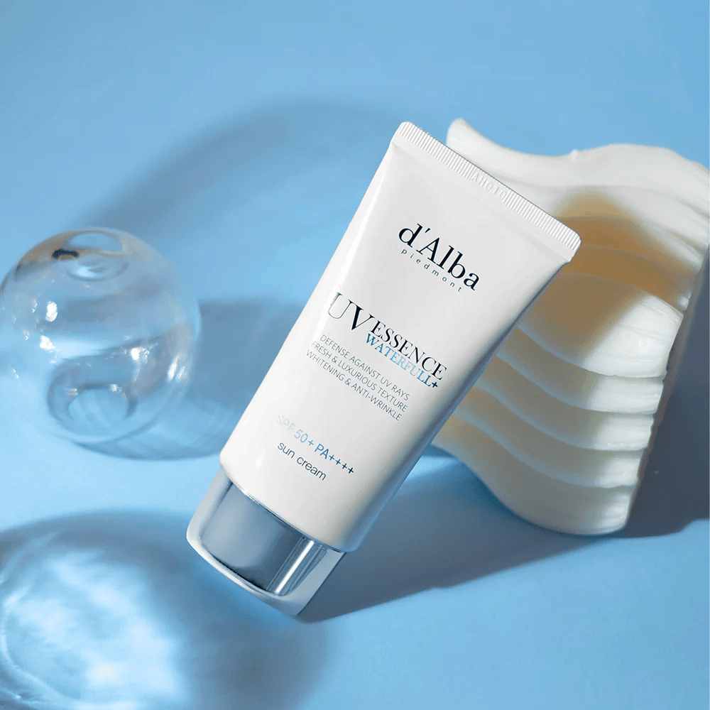 d'Alba UV Essence Waterfull+ Sun Cream so SPF 50+ PA++++ leží na modrom pozadí s odrazmi svetla a dekoratívnymi prvkami – zdôrazňuje svieže a hydratačné vlastnosti opaľovacieho krému.