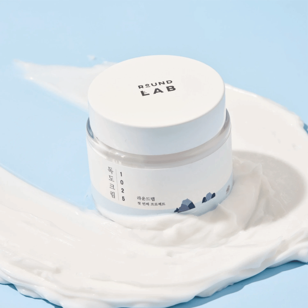 Pohár s Round Labs 1025 Dokdo Cream 80 ml, ktorý poskytuje hydratáciu, umiestnený na žiarivom modrom pozadí.