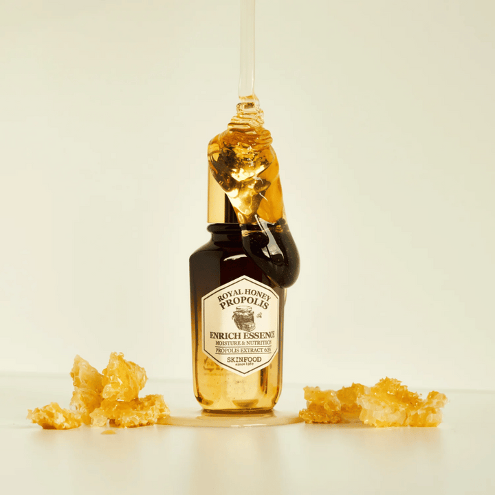Obrázok sklenenej fľaše s Royal Honey Propolis Enrich Essence od Skinfood. Veľká kvapka zlatej medu steká z fľaše so zlatým uzáverom. Okolo fľaše sú roztrúsené malé kúsky medového plástu na svetlom povrchu. Esencia je propagovaná so zameraním na vlhkosť a výživu, s 63% propolisového extraktu.