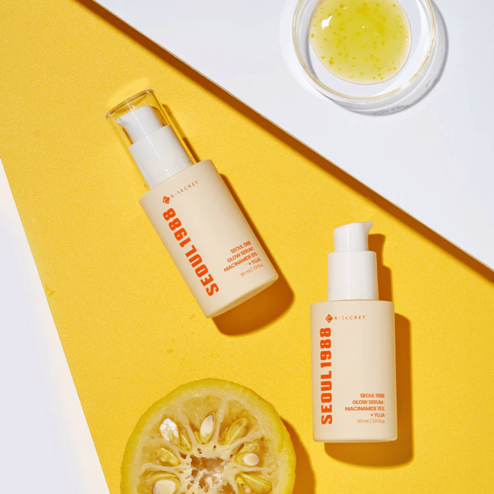 Dve fľaše Seoul 1988 Glow Serum umiestnené na žltom pozadí spolu s miskou so žltým sérom a polovičným citrusovým plodom.