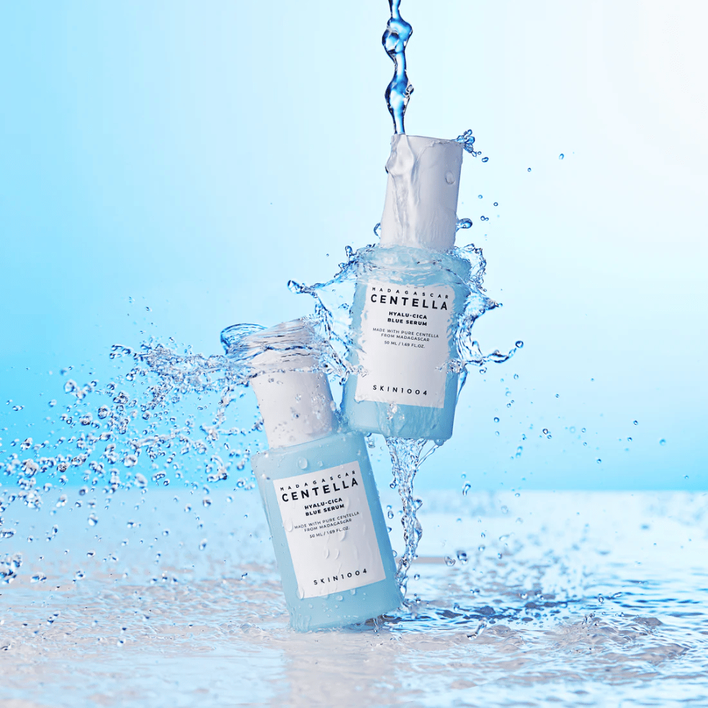 Dve fľaše SKIN1004 Madagascar Centella Hyalu-Cica Blue Serum sú zasiahnuté kaskádou vody na svetlomodrom pozadí.