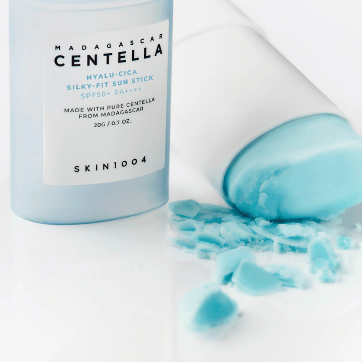 Tyčinka na opaľovanie s SPF50+ leží otvorená vedľa svojho vrchnáka, obsah čiastočne rozotretý na bielej ploche. Text na obale spomína ingrediencie z Madagaskaru.