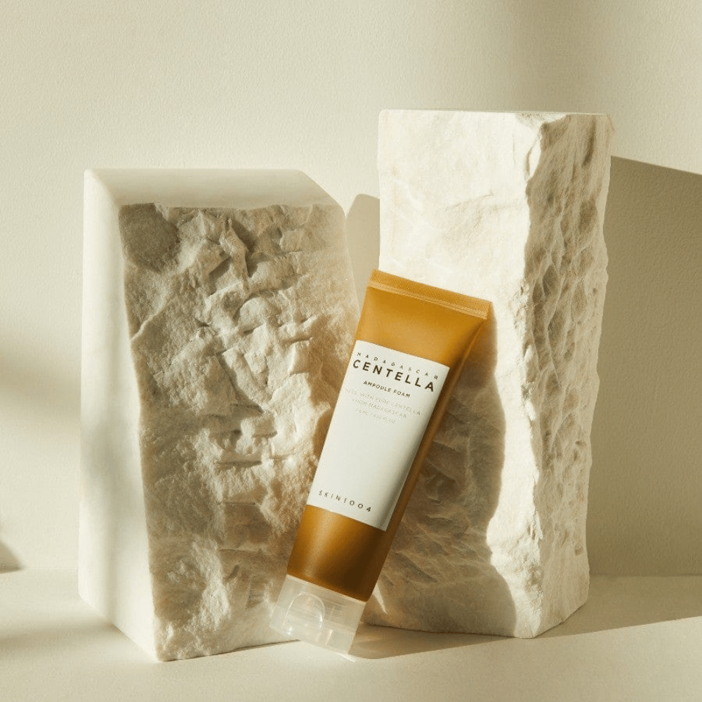 SKIN1004 Madagascar Centella Ampoule Foam stojaci proti svetlým kamenným blokom v prirodzenom slnečnom svetle, s jeho hnedobielym dizajnom tuby a elegantnou minimalizmou.