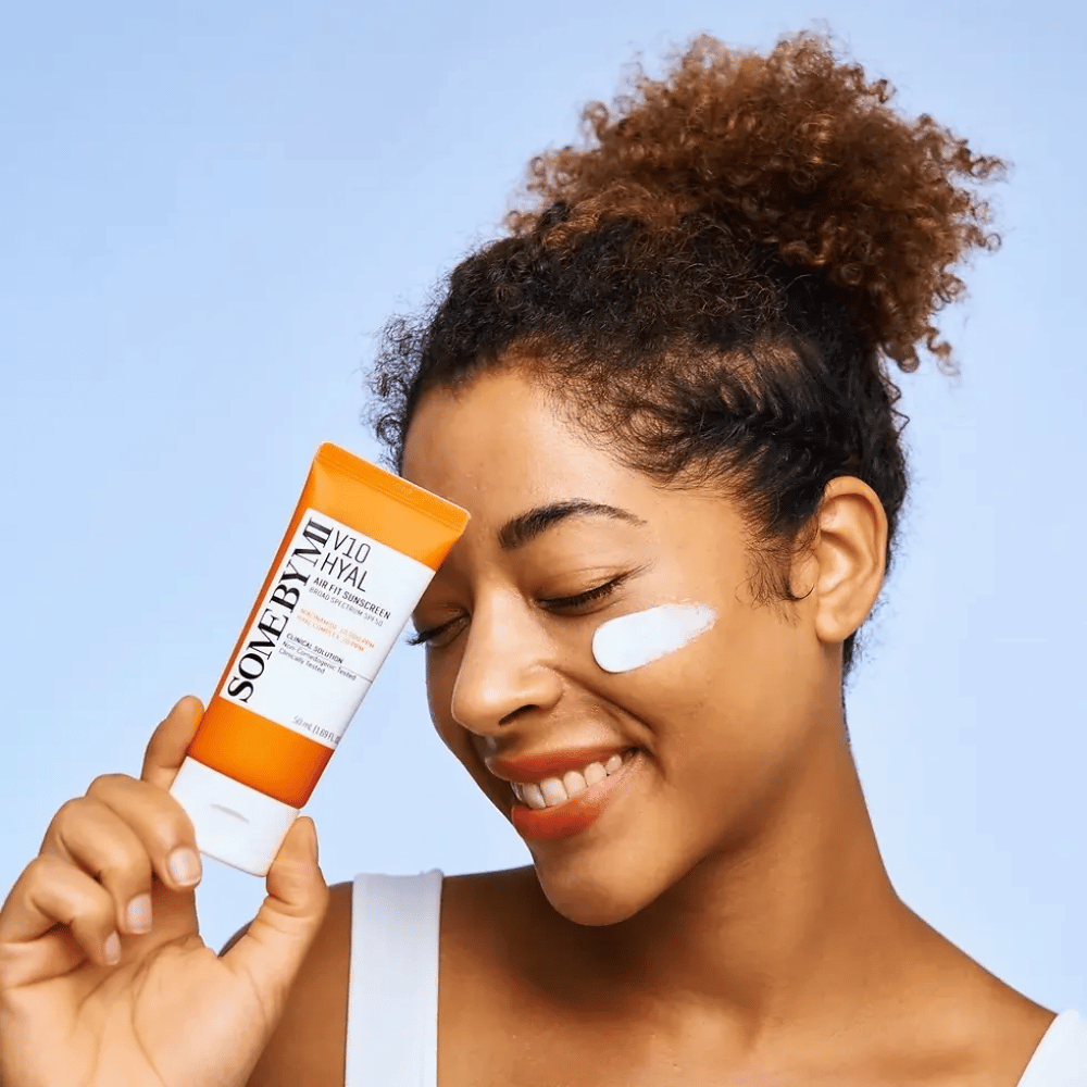 Žena drží tubu SOME BY MI V10 Hyal Air Fit Sunscreen a má bielu škvrnu opaľovacieho krému na líci. S úsmevom ukazuje produkt, čím jasne zdôrazňuje jeho použitie a výhody pre starostlivosť o pleť a ochranu pred slnkom.
