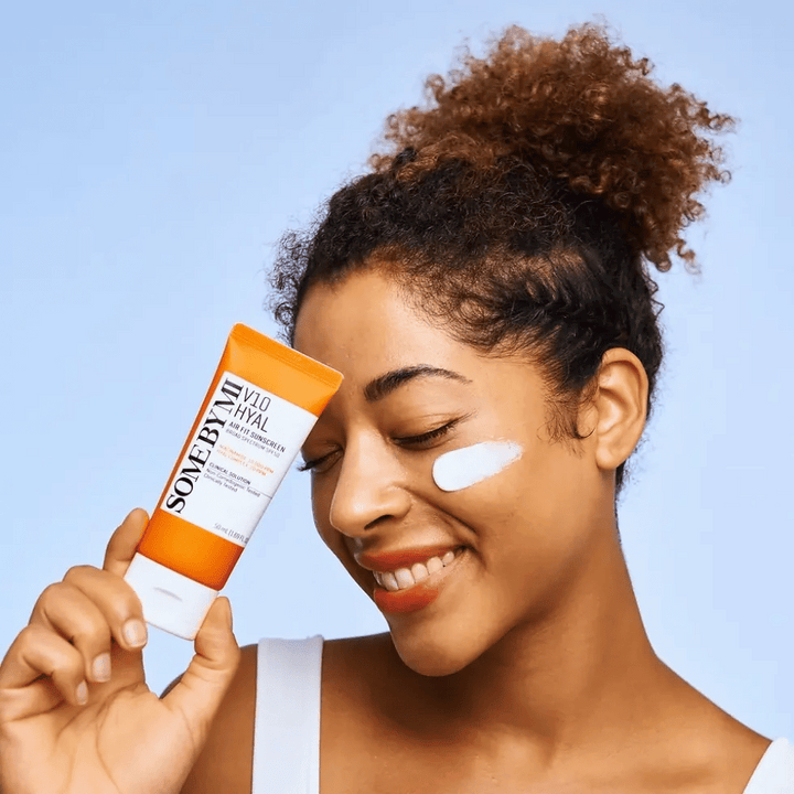 Žena drží tubu SOME BY MI V10 Hyal Air Fit Sunscreen a má bielu škvrnu opaľovacieho krému na líci. S úsmevom ukazuje produkt, čím jasne zdôrazňuje jeho použitie a výhody pre starostlivosť o pleť a ochranu pred slnkom.