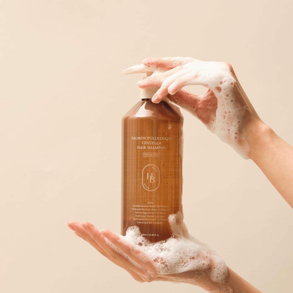 Heveblue Salmon Pullkeratin Centella Hair Shampoo v hnedej fľaši obklopenej penou. Produkt je držaný medzi dvoma rukami a zdôrazňuje čistiaci a ošetrujúci zážitok z vlasovej starostlivosti s bohatou penou.
