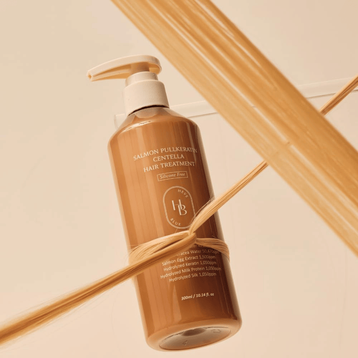 Heveblue Salmon Pullkeratin Centella Hair Treatment v hnedej fľaši obklopená prameňmi vlasov. Obrázok zdôrazňuje ošetrujúce vlastnosti vlasovej kúry so zameraním na lesk, silu a hodvábne jemné vlasy.