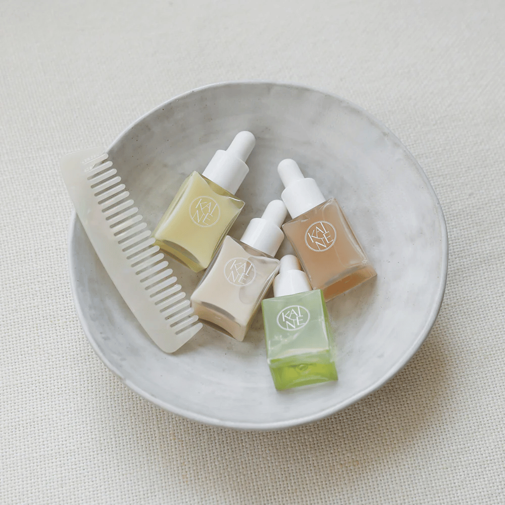 Štyri malé fľaštičky séra z KAINÉ Serum Discovery Kit ležia v bielej miske vedľa hrebeňa, s prirodzeným a pokojným pozadím v béžovom tóne.