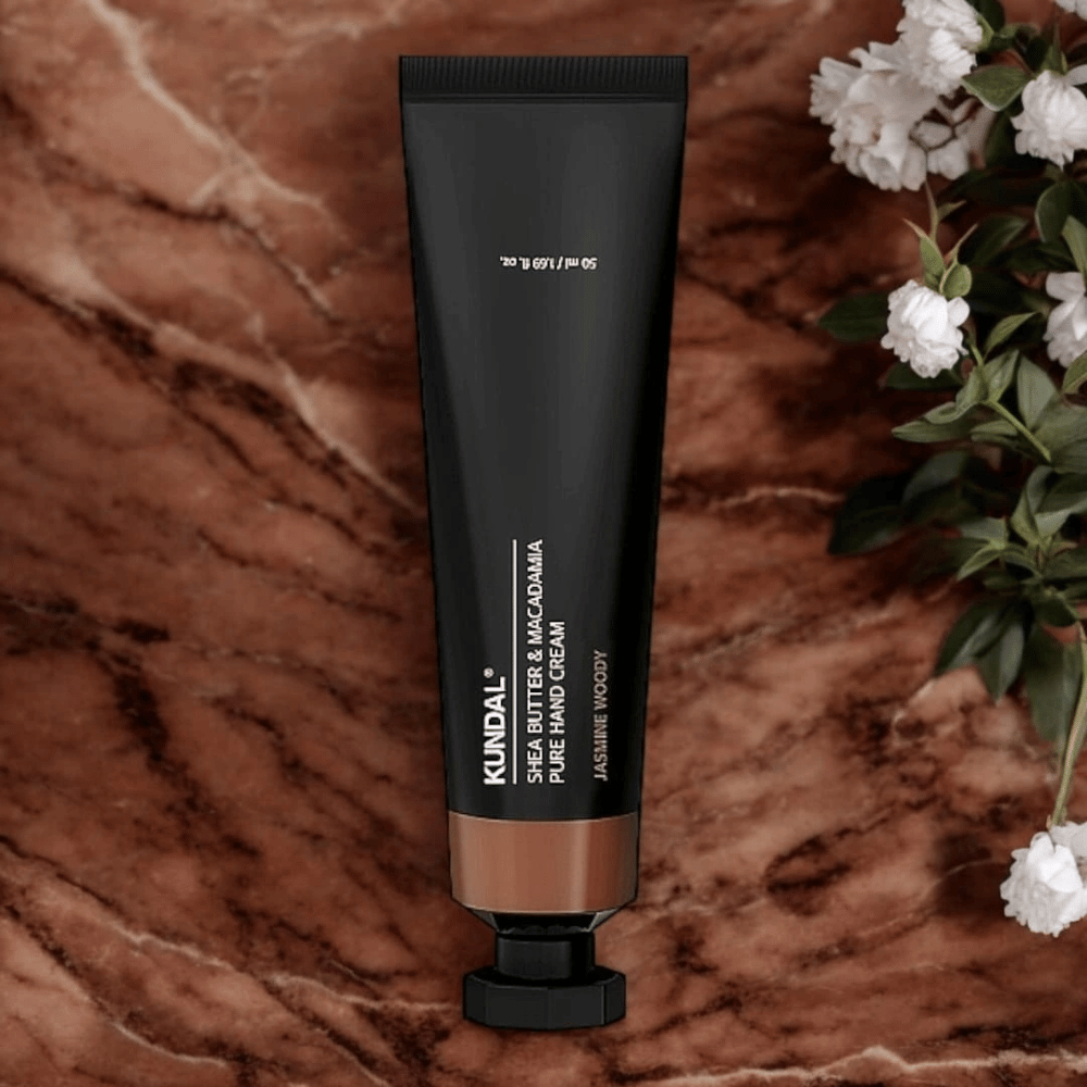 KUNDAL Shea Butter & Macadamia Pure Hand Cream Jasmine Woody umiestnený na mramore s kvetmi – výživný krém na ruky, ktorý ošetruje suché ruky a poskytuje elegantnú vôňu jazmínu a drevité tóny.