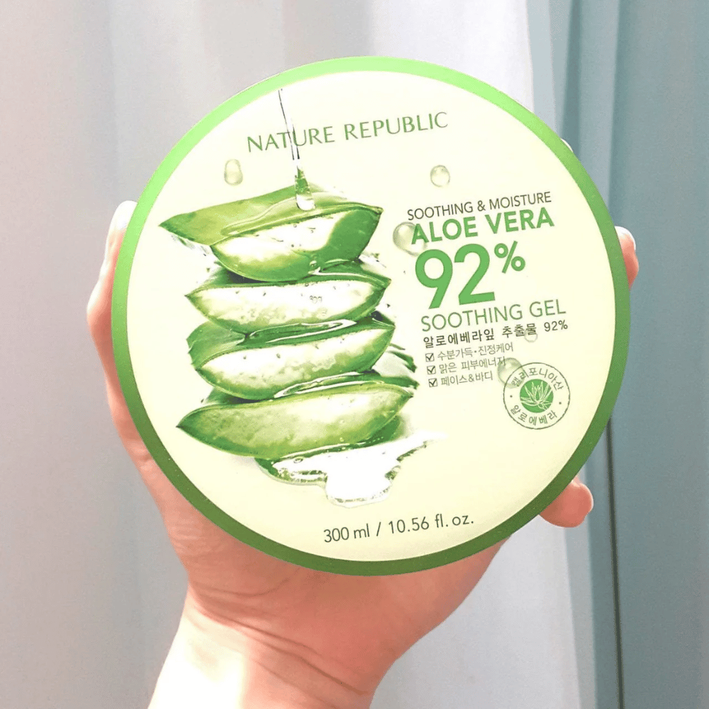 Ruka drží okrúhlu nádobu s Nature Republic Aloe Vera 92% Soothing Gel. Nádoba je zelená s obrázkom aloe vera a textom, ktorý popisuje jej upokojujúce a hydratačné vlastnosti. Obsah je 300 ml.