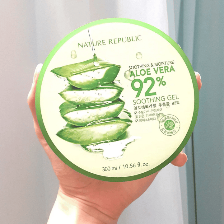 Ruka drží okrúhlu nádobu s Nature Republic Aloe Vera 92% Soothing Gel. Nádoba je zelená s obrázkom aloe vera a textom, ktorý popisuje jej upokojujúce a hydratačné vlastnosti. Obsah je 300 ml.