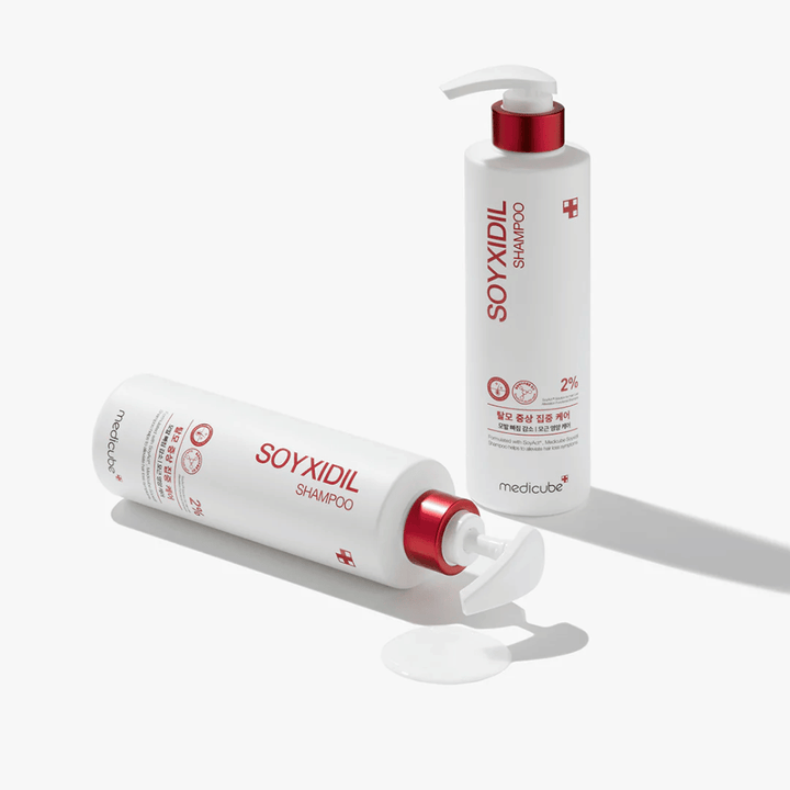 Dve fľaše Medicube Soyxidil Shampoo v bielom a červenom dizajne, jedna stojaca a jedna ležiaca s rozliatym šampónom vedľa.
