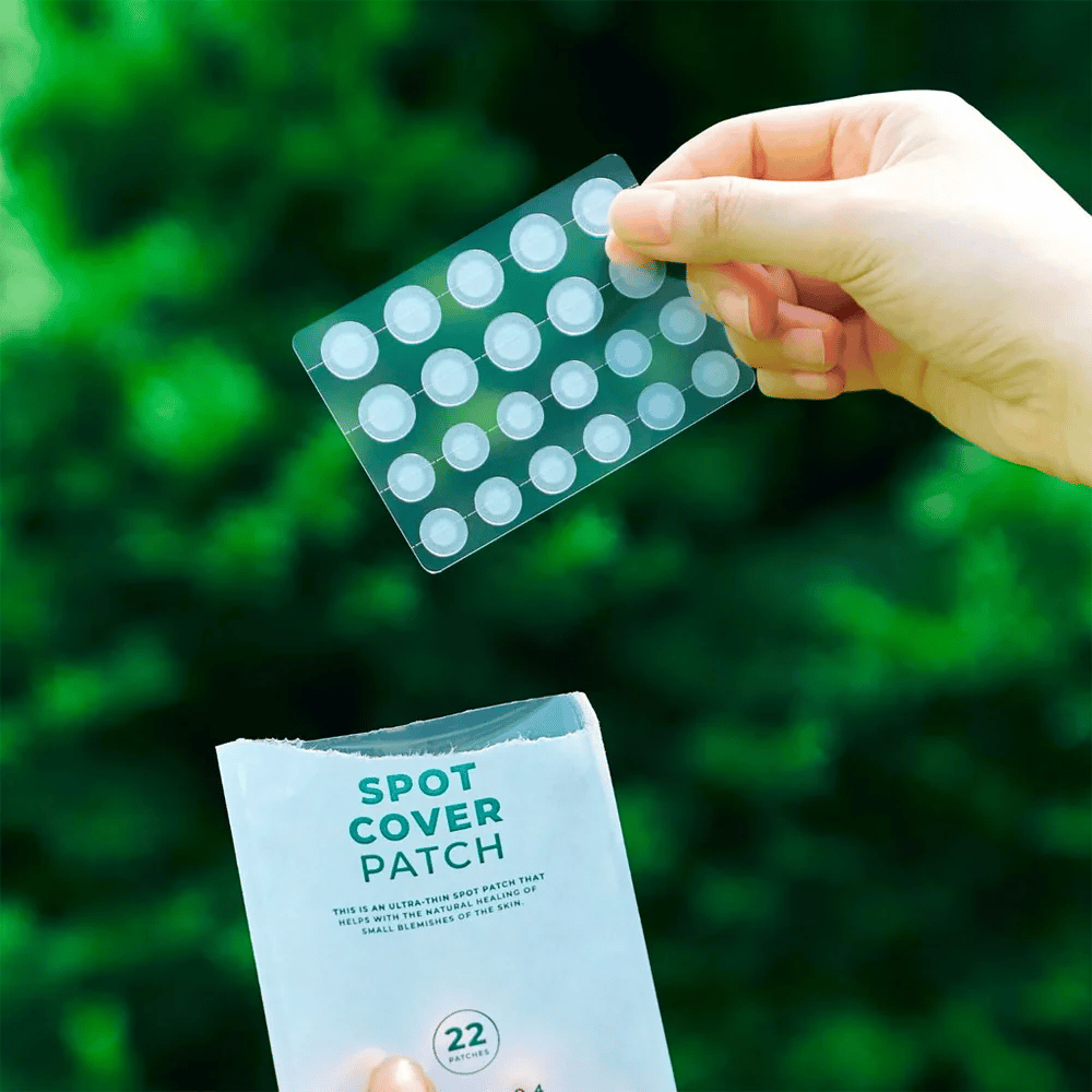 Ruka drží hárok s náplasťami Spot Cover Patches, ktoré sú priehľadné a zaoblené. V pozadí je rozmazané zelené prostredie. Balenie ukazuje časť balenia s textom "SPOT COVER PATCH" a informáciou, že produkt obsahuje 22 náplastí, ktoré pomáhajú liečiť malé oblasti pokožky.
